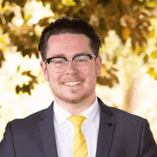 a man in a suit and tie is smiling for the camera while standing in front of a tree .