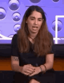 a woman with long hair and a tattoo on her arm is sitting in front of a television and making a funny face .