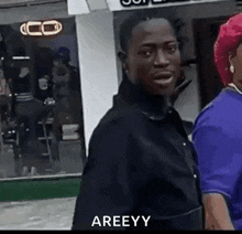 a man in a black jacket is standing next to a woman in a blue shirt and says areeyy .