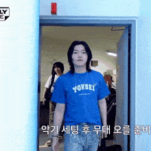a young man wearing a blue yonsei t-shirt stands in a doorway