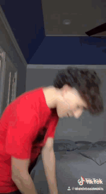 a young man in a red shirt is standing on a bed in a bedroom .