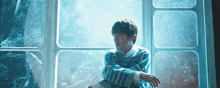 a young man in a blue striped sweater is sitting in front of a window