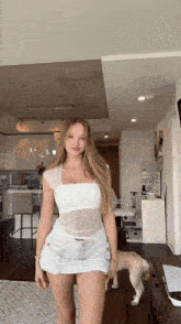 a woman in a white top and white shorts is standing in a living room with a dog standing behind her .