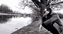 a man is smoking a cigarette while squatting next to a tree by a lake .