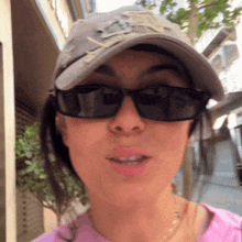 a woman wearing sunglasses a hat and a pink shirt looks at the camera