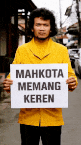 a man in a yellow jacket is holding a sign that says mahkota emang keren