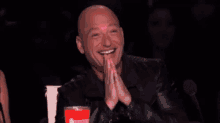 a bald man is sitting in front of a microphone with his hands folded in prayer .