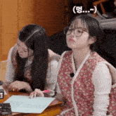 two girls are sitting at a table with one wearing glasses and a floral vest