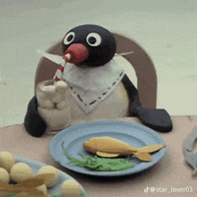 a stuffed penguin is sitting at a table with a plate of food and drinking from a straw .