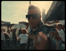 a man wearing sunglasses stands in front of a wall with graffiti that says rt