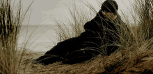 a person in a black coat is laying in the sand near tall grass
