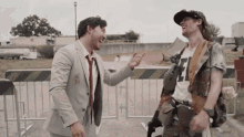 two men are standing next to each other in a field and laughing . one of the men is wearing a suit and tie .
