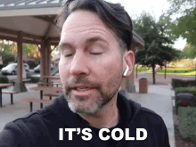 a man wearing ear buds is standing in a park with his eyes closed and says it 's cold .