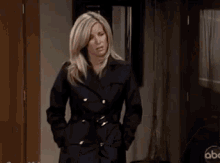 a woman in a black trench coat is standing in a hallway with her hands in her pockets .