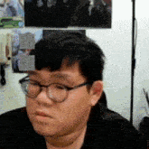 a man wearing glasses and a black shirt is sitting in front of a wall with pictures on it