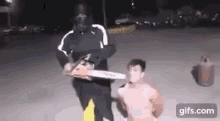 a man in a mask is holding a chainsaw in front of a man without a shirt .