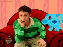 a man in a green and black striped shirt is sitting on a red couch next to a blue dog
