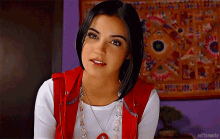 a woman wearing a red vest and a white shirt is standing in front of a wall with a tapestry on it