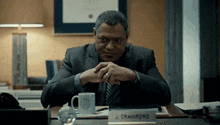 a man in a suit and tie sits at a desk with a name plate that says j crawford