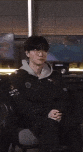 a man wearing glasses and a black jacket is sitting in a chair and clapping his hands .