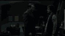 two men standing next to each other in a dark room with a clock on the wall
