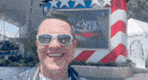 a man wearing sunglasses is smiling in front of a capitol fourth sign