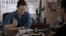 a man in a blue shirt is giving a thumbs up while sitting at a desk and talking to another man .