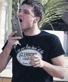 a man wearing a clermont lounge t-shirt is holding a cell phone in his mouth