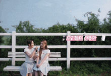 two women sit on a bench in front of a white fence with a sign that says ' 우리들 의 신붓한 '