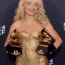 a woman in a gold dress holds up two grammy awards