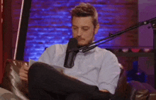 a man is sitting in a chair with his legs crossed in front of a microphone holding a piece of paper .