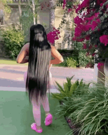 a woman with long black hair and pink shoes is standing in front of a bush with pink flowers .