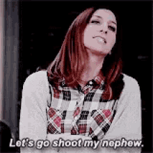 a woman in a plaid shirt is standing with her arms crossed and says `` let 's go shoot my nephew . ''