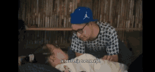 a man wearing a blue hat and glasses talks to a girl laying on a bed
