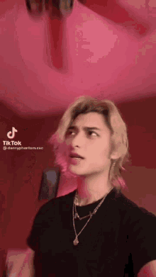 a young man with blonde hair is wearing a black shirt and a necklace and has a tik tok account .
