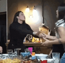 two women are fighting in a kitchen with a table full of plates and glasses