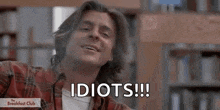 a young man in a plaid shirt is sitting in a library and says `` idiots '' .