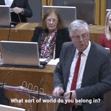 a man in a suit and tie stands at a podium with the words " what sort of world do you belong in " below him