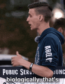 a man in a blue hoodie stands in front of a sign that says " public service biologically that 's "