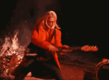 a man in an orange coat playing a guitar in front of a fire