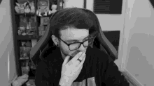 a man wearing glasses and a ring is sitting in a chair covering his mouth .