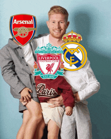 a man holds a child in front of a liverpool and real madrid emblem