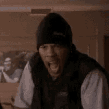 a man wearing a beanie and a vest is sitting in a room with the words `` the fuck '' written on his face .
