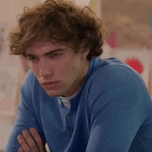 a young man with curly hair is wearing a blue sweater and earrings and looking at the camera .