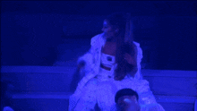 ariana grande is singing into a microphone while sitting on a stage in a blue light .