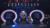 a man in a yellow suit stands in front of a large screen that says live
