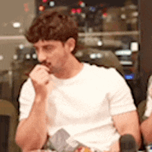 a man in a white t-shirt is sitting at a table with his hand on his chin .