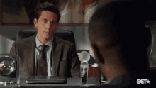 a man in a suit and tie sits at a desk with a bet logo on the bottom right