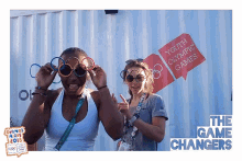 two women wearing sunglasses in front of a sign for youth olympic games