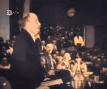 a man is giving a speech in front of a crowd with 24 ack written on the bottom right